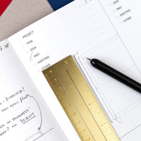 Brass Bookmark Ruler used in an opened book with a pen on a desk setting. 