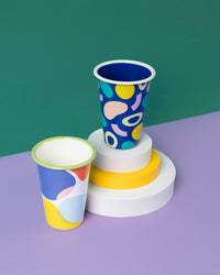 The Poketo x Crow Crayon Tumbler in Sea Glass next to the Tumbler in Outlines and Boulders on a green and purple background.